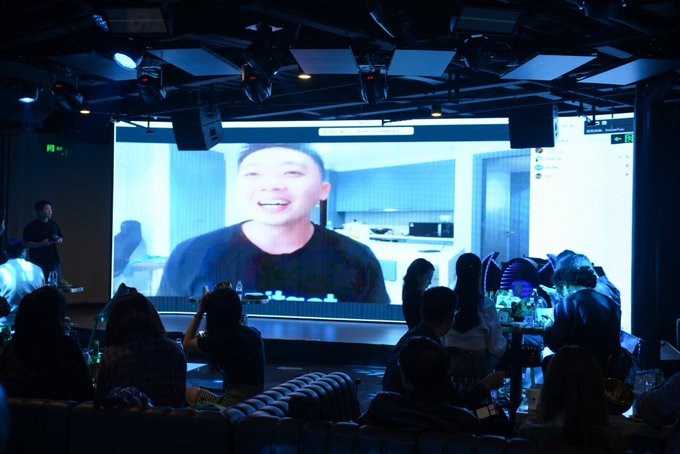 An audience in a dimly lit room watches a large screen displaying a smiling man during what appears to be a video call or live stream event.