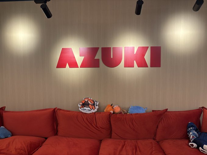 The image features a red couch with two stuffed animals sitting on top, including an orange elephant and a blue whale plushie. The couch is positioned against a wall with a large "AZZUKI" sign above it. The text in the image suggests that the cultural layer of Ethereum was invited to the HQ of one of the most legendary components of that said Ethereum culture.