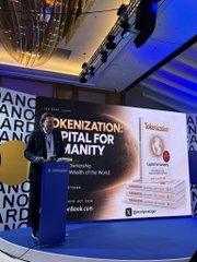 A man in a suit gives a presentation on a blue-lit stage in front of a screen displaying 'TOKENIZATION: CAPITAL FOR HUMANITY' and 'CBK', with Webull logos visible on the backdrop.
