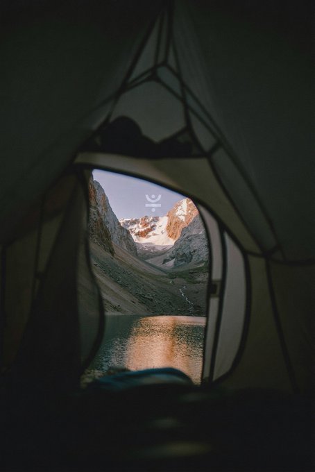 A view from inside a dark tent looks out through an opening to reveal a mountain lake reflecting snow-capped peaks at dusk.
