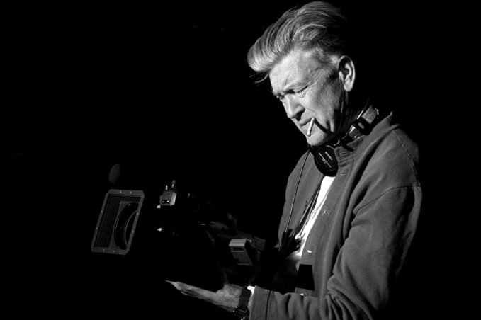 A black-and-white image of an older man with grey hair smiling while looking down at a professional camera, wearing headphones around his neck and a jacket over a t-shirt.