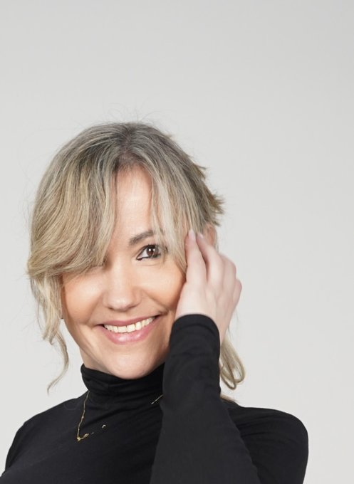A portrait of a smiling woman with blonde hair wearing a black turtleneck, resting her hand near her face against a light gray background.