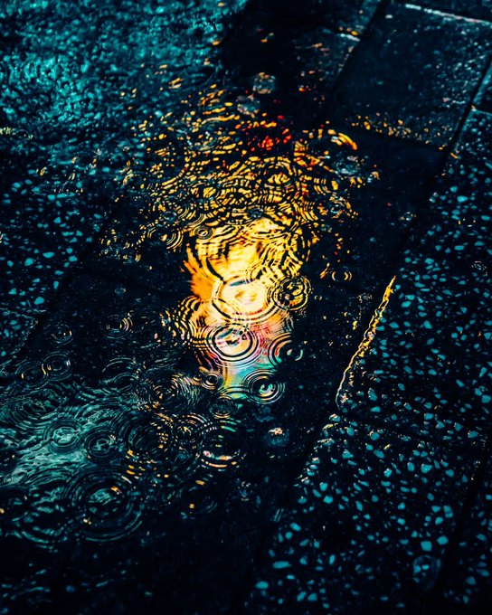 A close-up of a puddle on dark asphalt showing concentric ripples from falling rain, reflecting vibrant teal, orange, and golden colors from nearby lights.