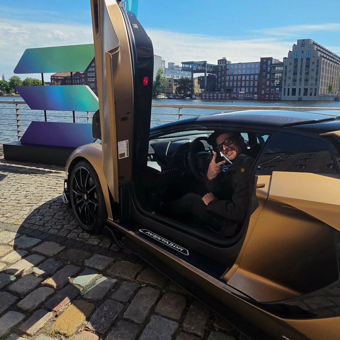A man wearing sunglasses and a black jacket sits in the driver's seat of a matte gold Lamborghini Aventador with its scissor door open, parked on a cobblestone waterfront with city buildings in the background.