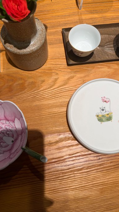 A wooden table set with a ceramic vase holding a red flower, a small teacup on a tray, and decorative plates including one featuring a cat illustration and another with a pink peony design.