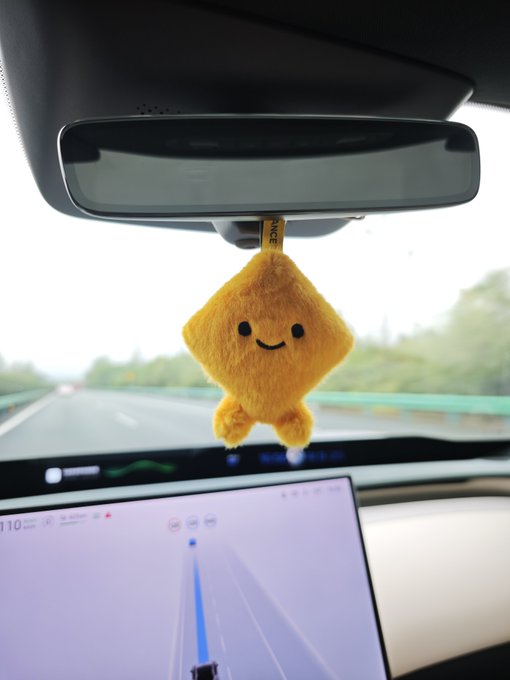 A small, yellow, smiling plush character hangs from a car's rearview mirror while driving on a highway, with the vehicle's navigation screen visible below.