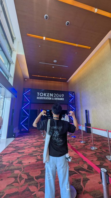 A person viewed from behind wearing a black t-shirt and gray pants, taking a photo of the 'TOKEN2049 REGISTRATION & ENTRANCE' signage inside a large convention hall with red patterned carpet.