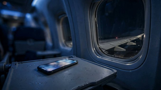 View from inside an airplane at night, showing the wing through the window and a smartphone resting on the tray table.
