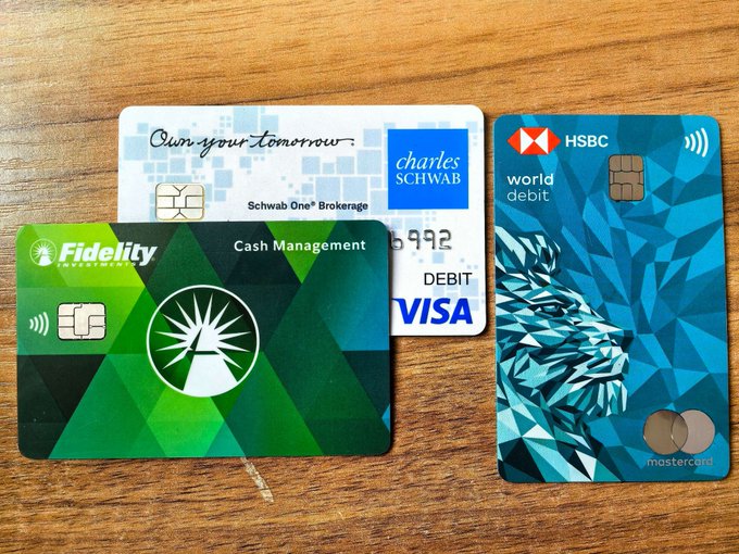 Three bank cards arranged on a wooden surface: a green Fidelity Cash Management card, a white Charles Schwab Visa Debit card, and a blue HSBC World Debit Mastercard with a lion design.