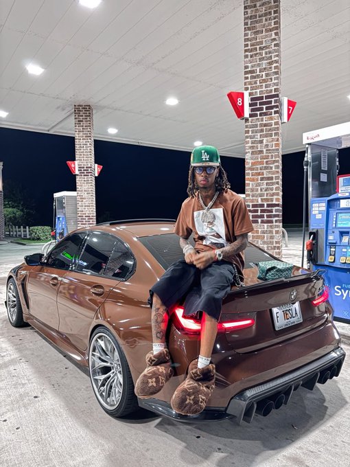 A man in a green LA cap and brown graphic tee sits on the trunk of a brown BMW at a gas station at night, posing for a flex photo to accompany his "Good morning web3 Gm CT" greeting.