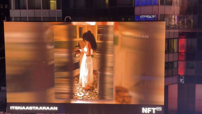 A large outdoor billboard in a city at night displays an artwork of a woman in a white dress with the text 'ITSNAASTAARAAN' and an 'NFT' logo.