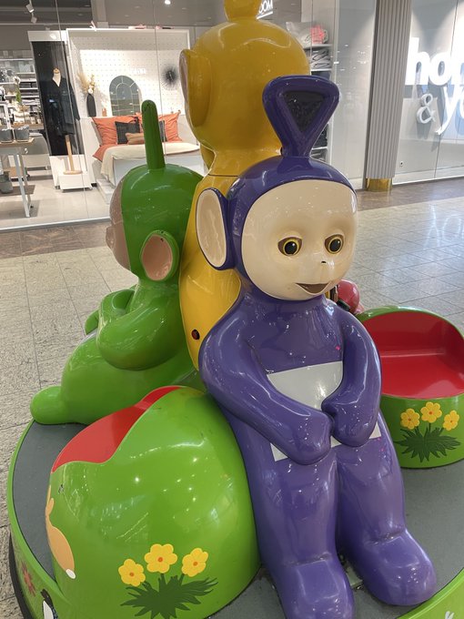 A colorful statue featuring Teletubbies characters (Tinky Winky in purple, Dipsy in green, and Laa-Laa in yellow) sitting together in what appears to be a shopping mall or store display.
