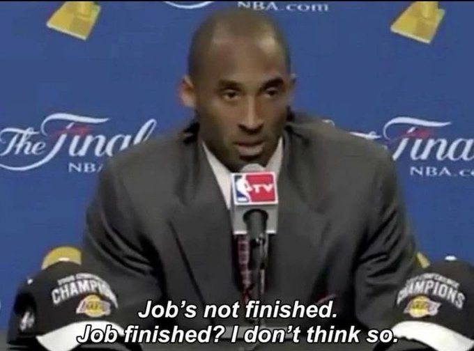 Kobe Bryant sits at a post-game press conference with championship hats beside him and "The Finals NBA" backdrop behind, captioned "Job's not finished. Job finished? I don't think so." The image visually reinforces the post's theme of relentless perseverance, echoing Bryant's iconic mindset that the pursuit of excellence never truly ends.