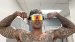 A heavily tattooed man wearing a pink beanie and reflective sunglasses flexes his biceps while taking a mirror selfie.