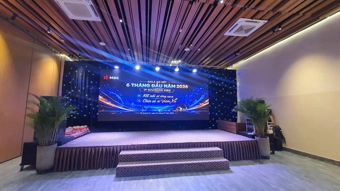 An event stage with a large screen displaying "GALA MỪNG 6 THÁNG ĐẦU NĂM 2026" (MBS), featuring decorative lighting and potted palm plants on either side of the stage.