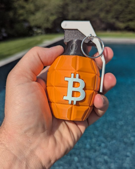A hand holding an orange grenade-shaped keychain featuring a white Bitcoin (₿) symbol, photographed outdoors near a swimming pool.