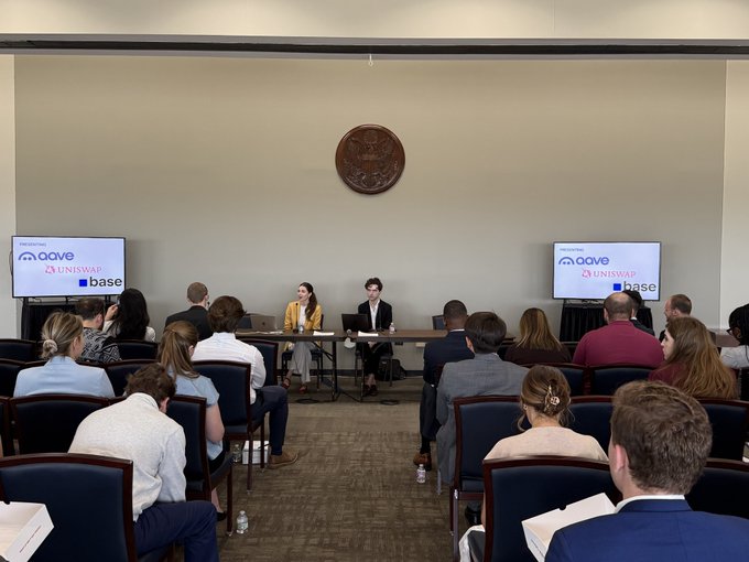 The image shows a group of people sitting in chairs around a table, with some individuals wearing suits. They are gathered together for a meeting or presentation related to cryptocurrency and blockchain technology. The room appears to be a conference room or a similar setting designed for such events. The attendees seem engaged and attentive as they listen to the speaker who is talking about DeFi protocols and their potential applications in the future.