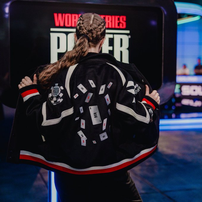A person with a long braided ponytail stands with their back to the camera, displaying a black jacket covered in small cards or stickers, with the words 'WORK' and 'POWER' visible on a screen in the background.