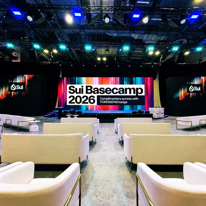 An empty event venue with rows of white chairs facing a stage displaying 'Sui Basecamp 2026 - Complimentary access with TOKEN2049 badge' on a colorful LED backdrop, with 'Sui' logos visible on side screens.