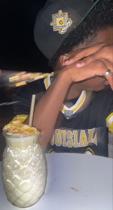 A person wearing a black baseball cap and a black 'LOUISIANA' jersey sits at a table with their hands covering their face, with a pineapple-shaped drink in the foreground.