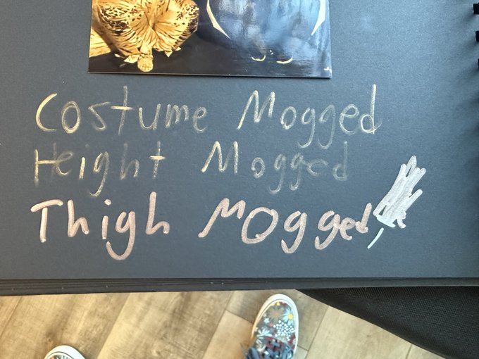 A chalkboard with handwritten text reading 'Costume Mogged, Height Mogged, Thigh Mogged' with scribbled-out text, with a person's feet in colorful patterned shoes visible at the bottom.