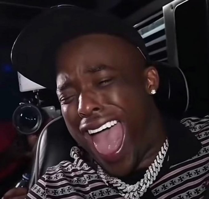 A close-up of a man wearing a black cap, diamond earrings, and a diamond chain necklace, smiling widely with his mouth open.