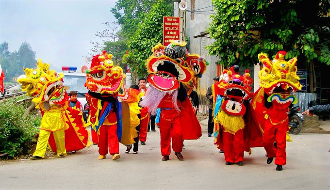 A vibrant street procession featuring performers in colorful traditional lion and dragon dance costumes, including large yellow and red lion heads and an elaborate dragon figure with a colorful mane, being carried and danced through a street with onlookers.