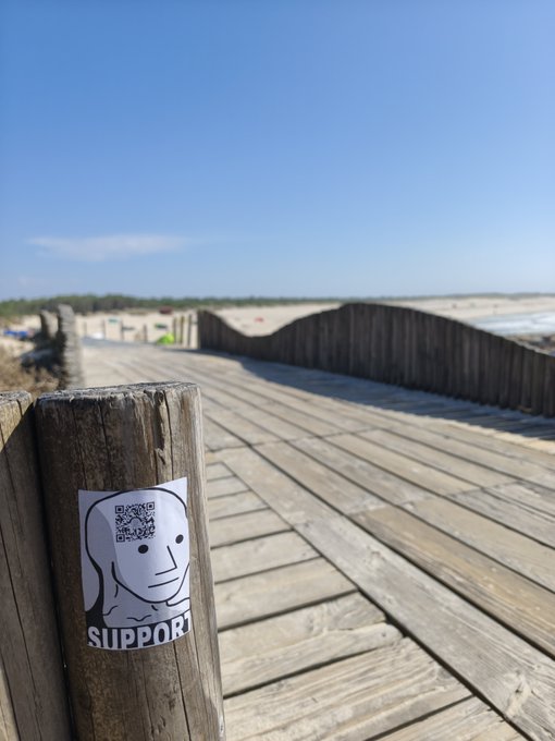 A wooden boardwalk leads toward a beach under a clear blue sky, with a sticker featuring a cartoon face and the word 'SUPPORT' attached to a wooden post in the foreground.