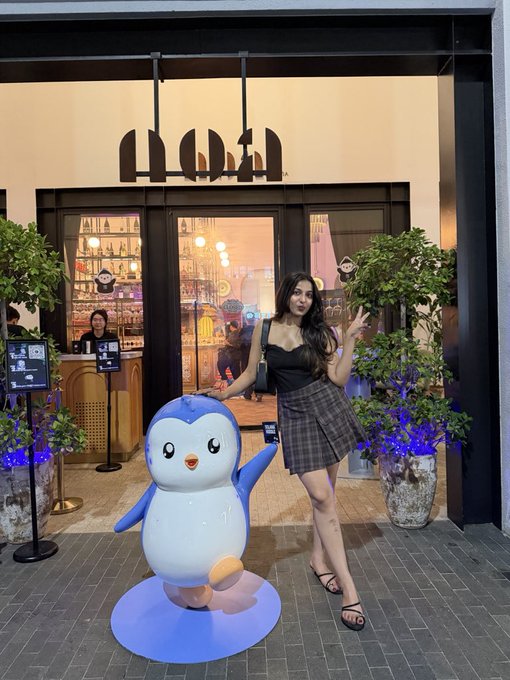 A woman poses beside a blue and white penguin mascot at the entrance of "hud" (Huddle), with the lit interior visible behind her, capturing the social vibe of the Pudgy Penguins × Solana meetup during TOKEN2049 in Singapore.