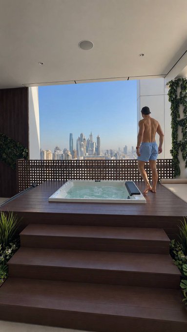 A shirtless man in blue shorts stands beside a private rooftop jacuzzi on a wooden deck, gazing out at a sweeping skyline view from his luxury penthouse. The image visually embodies the post's narrative of humble beginnings transformed into extraordinary financial success through perseverance in crypto trading.