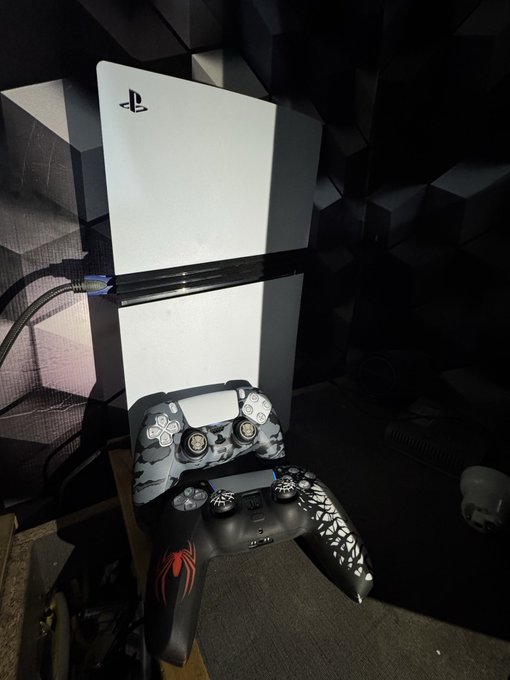 A PlayStation 5 console stands vertically with two custom DualSense controllers—one in gray/white camouflage and one in black with red flame and white graphic designs—resting against it.