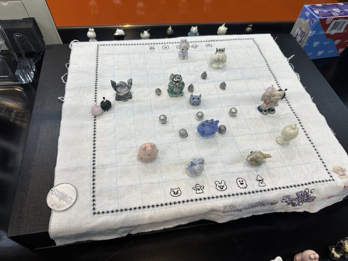 A small cloth game board with small figurines, shells, and coins arranged on a grid pattern on a dark table.
