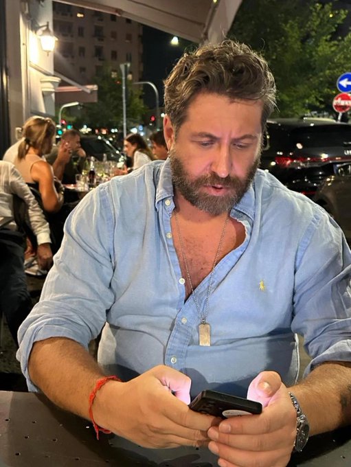 A bearded man in an unbuttoned denim shirt sits at an outdoor café at night, intently looking at his phone while a red string bracelet is visible on his wrist. In the background, a "Sens Unico" street sign can be seen, suggesting the scene takes place in Italy—perhaps reinforcing the "#bearish" caption's playful tone as the man appears focused on negative news or market trends on his screen.