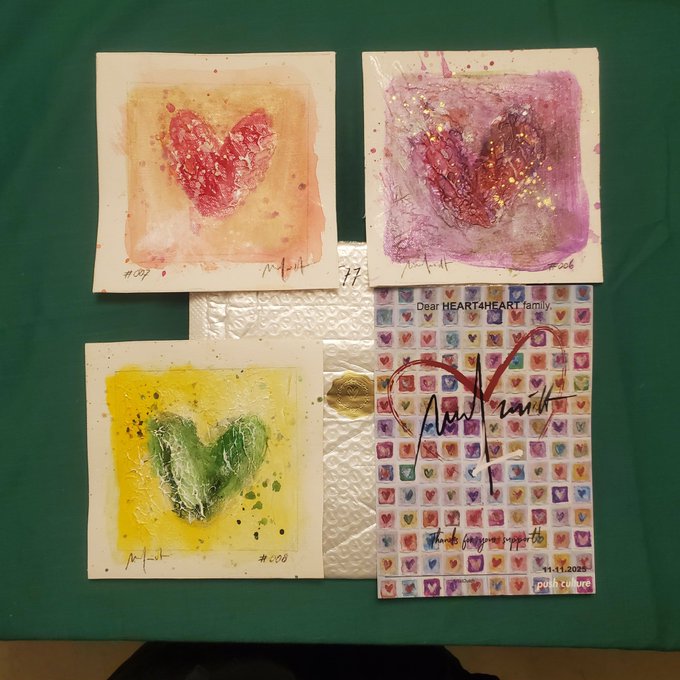 Four small canvas artworks featuring colorful painted hearts (red, purple, green/yellow) arranged on a green surface alongside a signed card reading 'Dear HEART4HEART family' dated 11.11.2023.