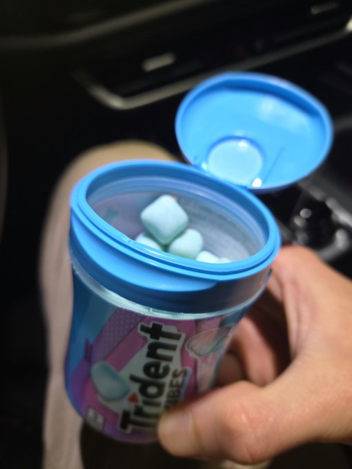 A hand holds an open container of Trident Vibes chewing gum, revealing light blue mints inside, inside a vehicle.