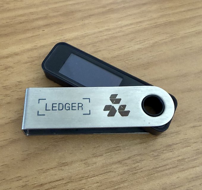 A photograph of a Ledger hardware cryptocurrency wallet lying open on a wooden surface, showing the silver metal casing with the "LEDGER" logo and the black device with its screen visible.