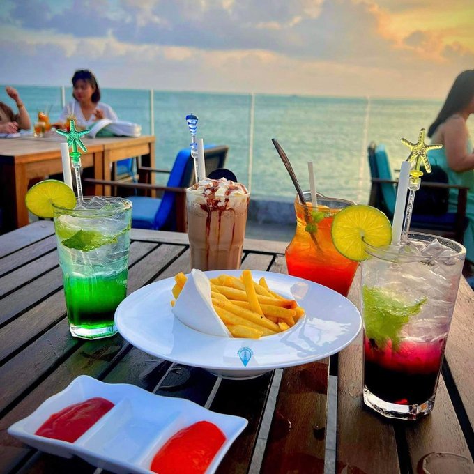 A wooden table set with colorful cocktails garnished with lime slices, a plate of french fries, and small dishes of red sauce, with people sitting by the ocean in the background.