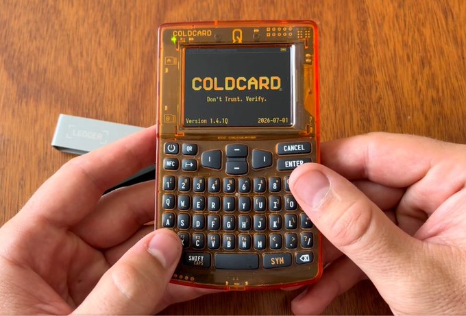 A person holds a transparent orange COLDCARD hardware wallet displaying 'Don't Trust. Verify. Version 1.4.10 2026-07-01' on its screen, with a Ledger device resting on a wooden table in the background.