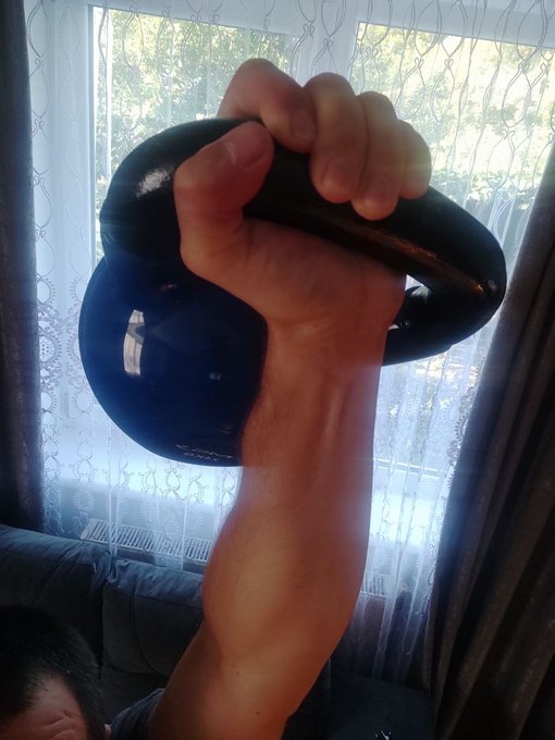 A close-up photograph of a muscular hand gripping a blue kettlebell in front of a window with sheer white curtains.