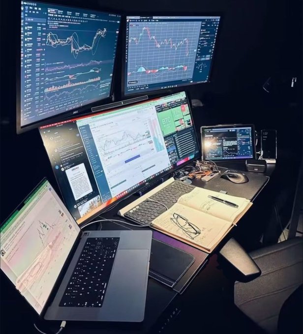 A trader's multi-monitor workstation glows in a dimly lit room, featuring two large overhead displays showing crypto candlestick charts with green and red price movements, a central monitor displaying a "BTC" Bitcoin dashboard with a $10,488.18 price tag and accompanying charts, and a tablet on the right running TradingView with additional market data, while an open MacBook in the foreground displays trading analytics alongside a notebook filled with handwritten notes and a pair of reading glasses, embodying the caption's message of trading as a self-built "money printer" that rewards those who master the analytical craft across multiple screens.