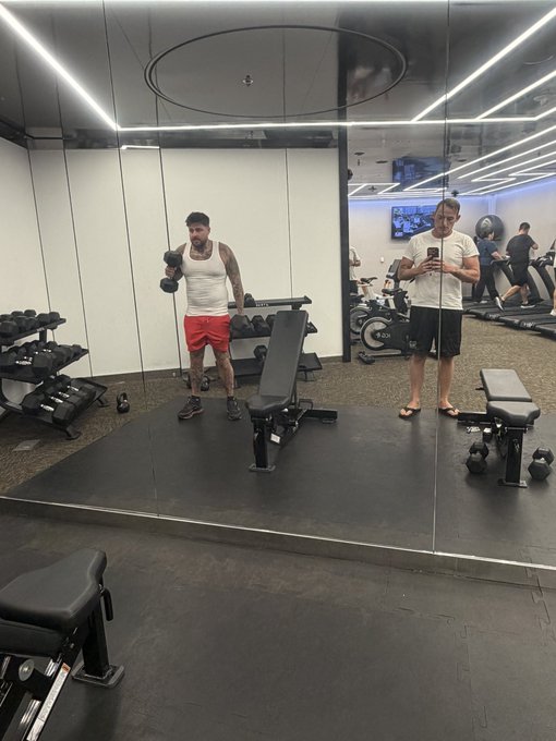A man performs a single-arm dumbbell lift in a gym mirror, while another man films him on his phone. The scene reflects a fitness-focused post tagged with @AscendXRP, suggesting a health and wellness promotion tied to the XRP community.