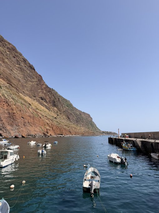 A small harbor filled with anchored boats sits nestled against a steep, reddish-brown cliff under a clear blue sky, with a stone pier extending into the calm water. The serene coastal scene creates a relaxed morning atmosphere that pairs well with the post's sunny greeting and invitation to share a Solana (SOL) crypto address.