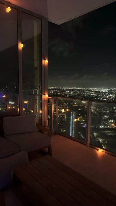 A nighttime view from a high-rise balcony shows a sprawling city skyline illuminated below, with a cozy seating area featuring a cushioned chair, wooden coffee table, and warm string lights framing the scene. The caption "GN frens, logging off" suggests the poster is signing off for the night, with the image capturing the tranquil, end-of-evening atmosphere from their apartment.