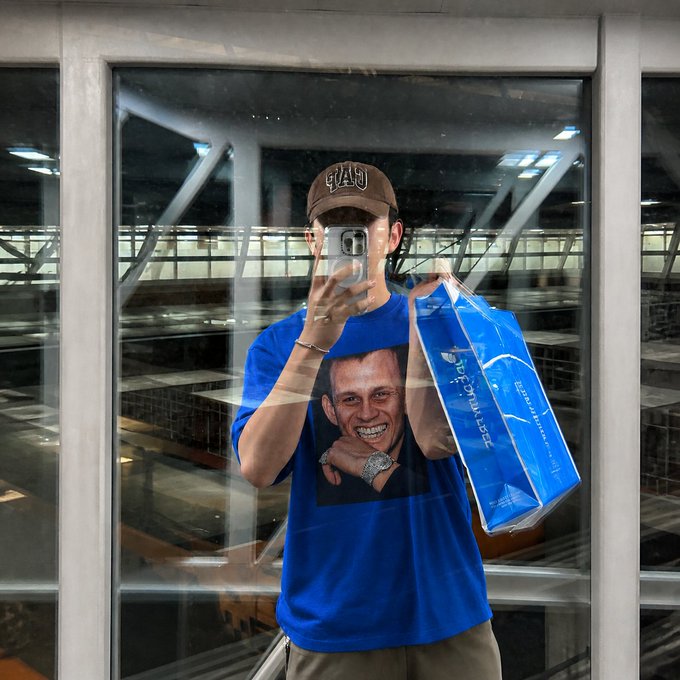 A person wearing a brown "GAP" cap takes a mirror selfie in a blue t-shirt featuring Vitalik Buterin's face, while holding a "Buterin.com" branded bag, fully embracing their Vitalik fanboy status.