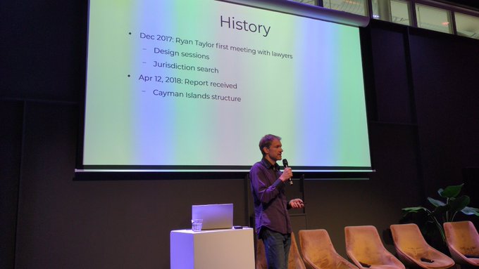 A man stands presenting in front of a projection screen titled 'History' that lists dates including 'Dec 2017: Ryan Taylor first meeting with lawyers' and 'Apr 12, 2018: Report received'.