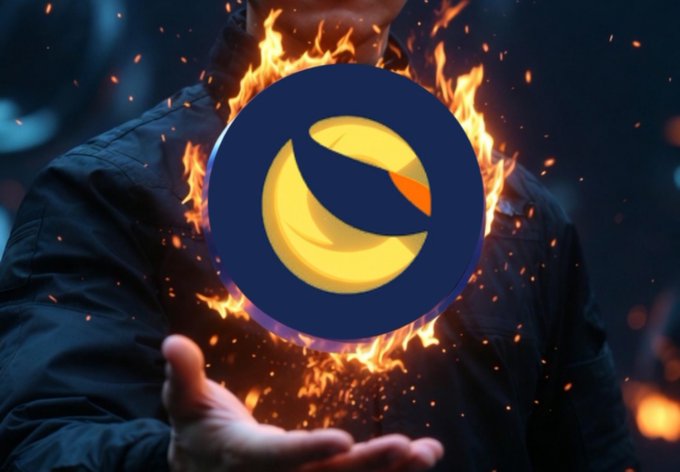 The LUNA/Terra cryptocurrency logo engulfed in flames being held in a person's hand, symbolizing the collapse or burning of the Terra Luna coin.