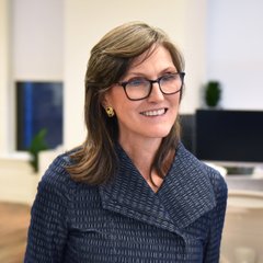 A woman with shoulder-length brown hair, wearing glasses and a navy blue textured blazer, smiles warmly in an office setting with blurred computer monitors in the background. The image accompanies a quote from Cathie Wood, the well-known investor and founder of ARK Invest, discussing her bullish outlook on blockchain technology.