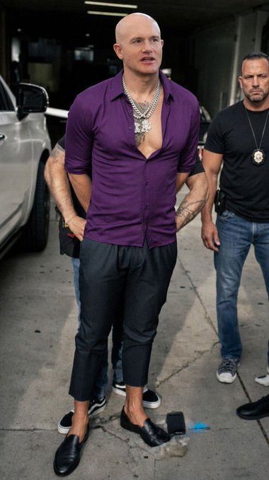A bald man wearing a purple short-sleeved button-up shirt, black pants, loafers, and prominent silver jewelry stands on a sidewalk next to a man in a black t-shirt who appears to be security or police.