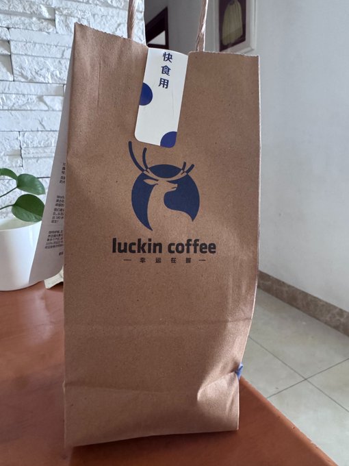 A brown paper bag from Luckin Coffee with the brand's deer logo and Chinese characters '快食用' on the tag, sitting on a table next to a potted plant.