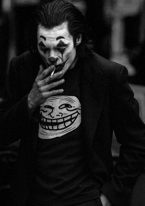 A black and white photo shows a man with clown makeup, slicked back hair, and a suit smoking a cigarette while wearing a t-shirt featuring a Trolls meme face. The image pairs with the post's joke about being "trolls," visually reinforcing the humor through the troll face graphic and the Joker-style clown aesthetic.
